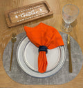 Amber Harvest table setting with grey oval placemat, orange linen napkin, and navy napkin ring